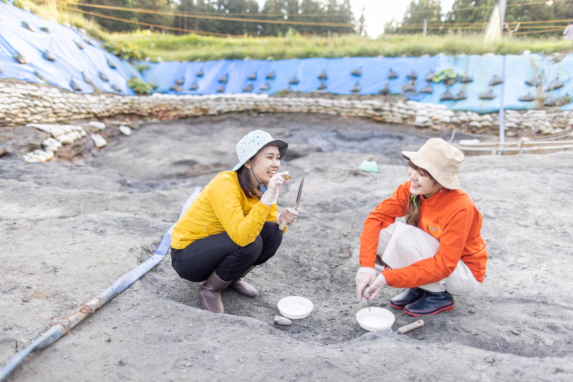 砂浜で帽子をかぶった2人の女性がしゃがみ、スコップで作業している写真
