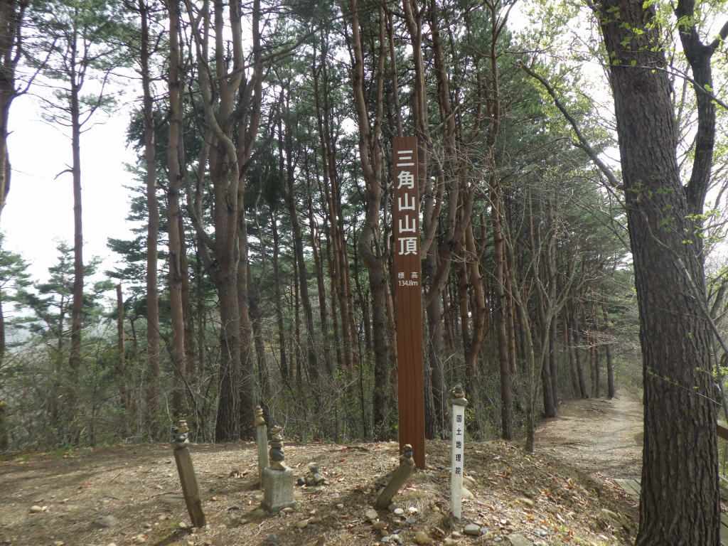 「三角山山頂」と書かれた表式が立っている森の中の写真