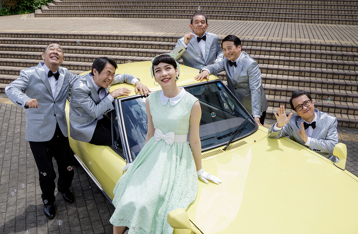 黄色の車の周りに男性5人が集まっており車体の前方に女性が座っている写真