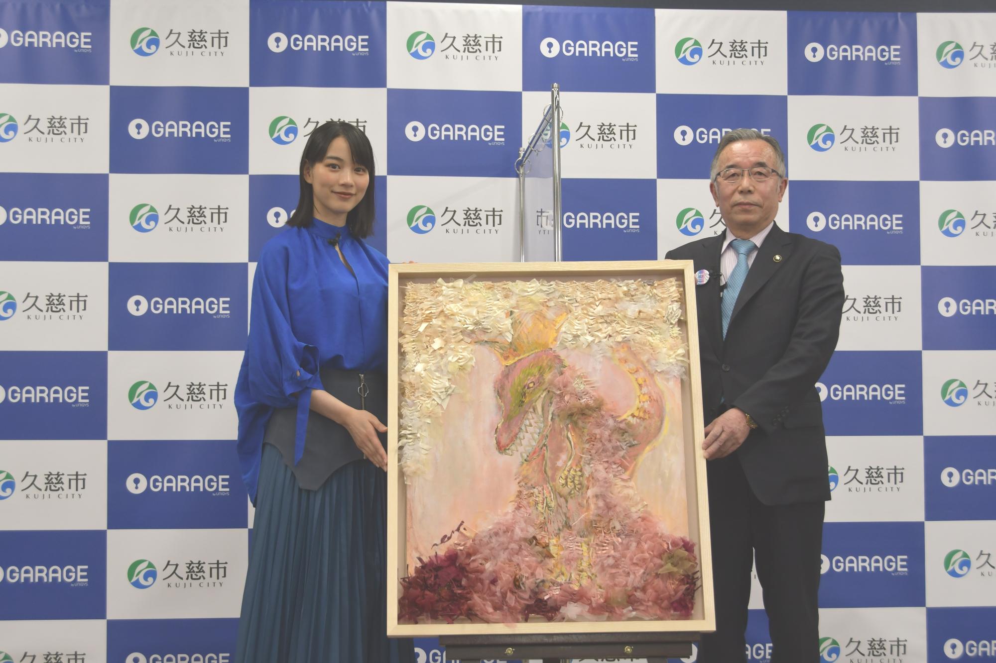 若い女性とスーツ姿の男性が左右に並び、1枚の大きな絵を持っている写真