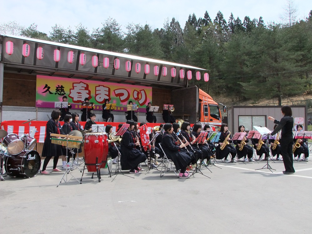 久慈春まつりと書かれた幕を背景に様々な楽器で演奏している人たちの写真