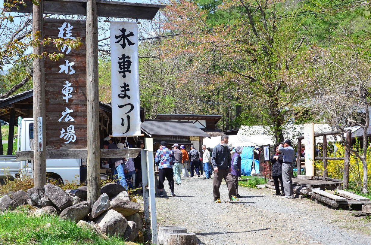 水車まつりや桂の水車広場と書かれた看板の奥にある広場に人が集まっている写真