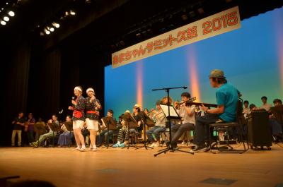 あまちゃんサミット久慈2015の幕を背景に踊りや演奏をしている人たちの写真