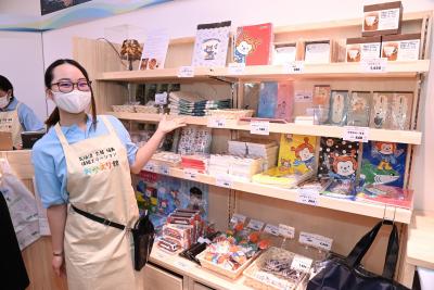 店の棚にグッズが並んでいるのをエプロン姿の女性が片手を差し出して示している写真