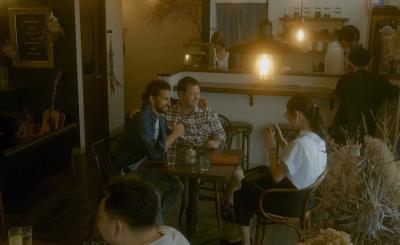 ランプの吊るされた店内で椅子に座って肩を組んでいる男性2人をカメラで撮影している女性の写真