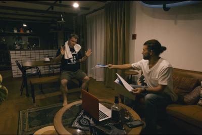 ソファに座っている男性が方にタオルを巻いている男性に紙を渡している写真