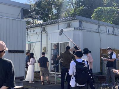プレハブ小屋の周りで撮影用のマイクなどを持った人たちが集まっている写真