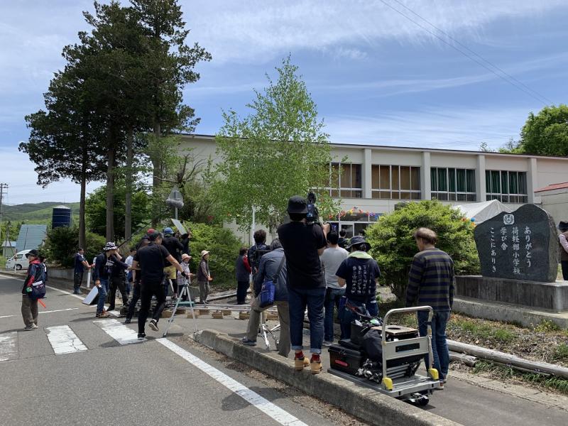 複数の人たちが集まっている学校の入り口でカメラで撮影をしている写真