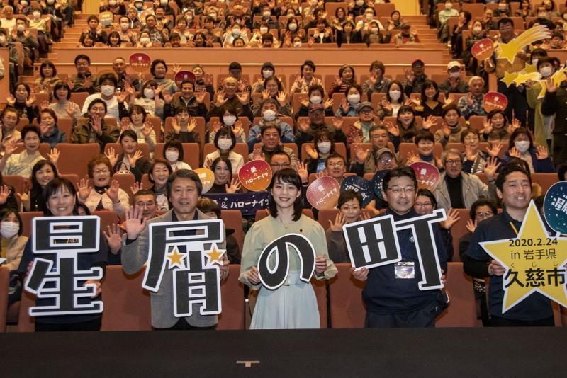 映画館の中で席に人が座っている前で映画のキャストが映画タイトルのパネルを持って笑っている写真