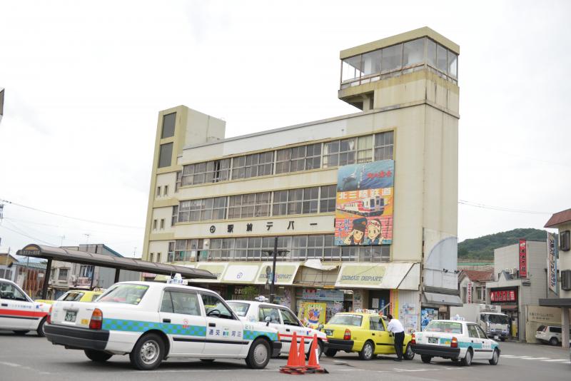 駅前デパートと書かれた4階建ての建物の前にタクシーが複数台停まっている写真