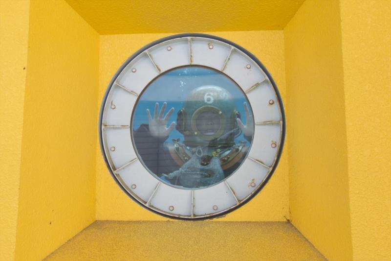 黄色の壁に設置された耐水ガラスの向こうで潜水服を着た人が手を振っている写真