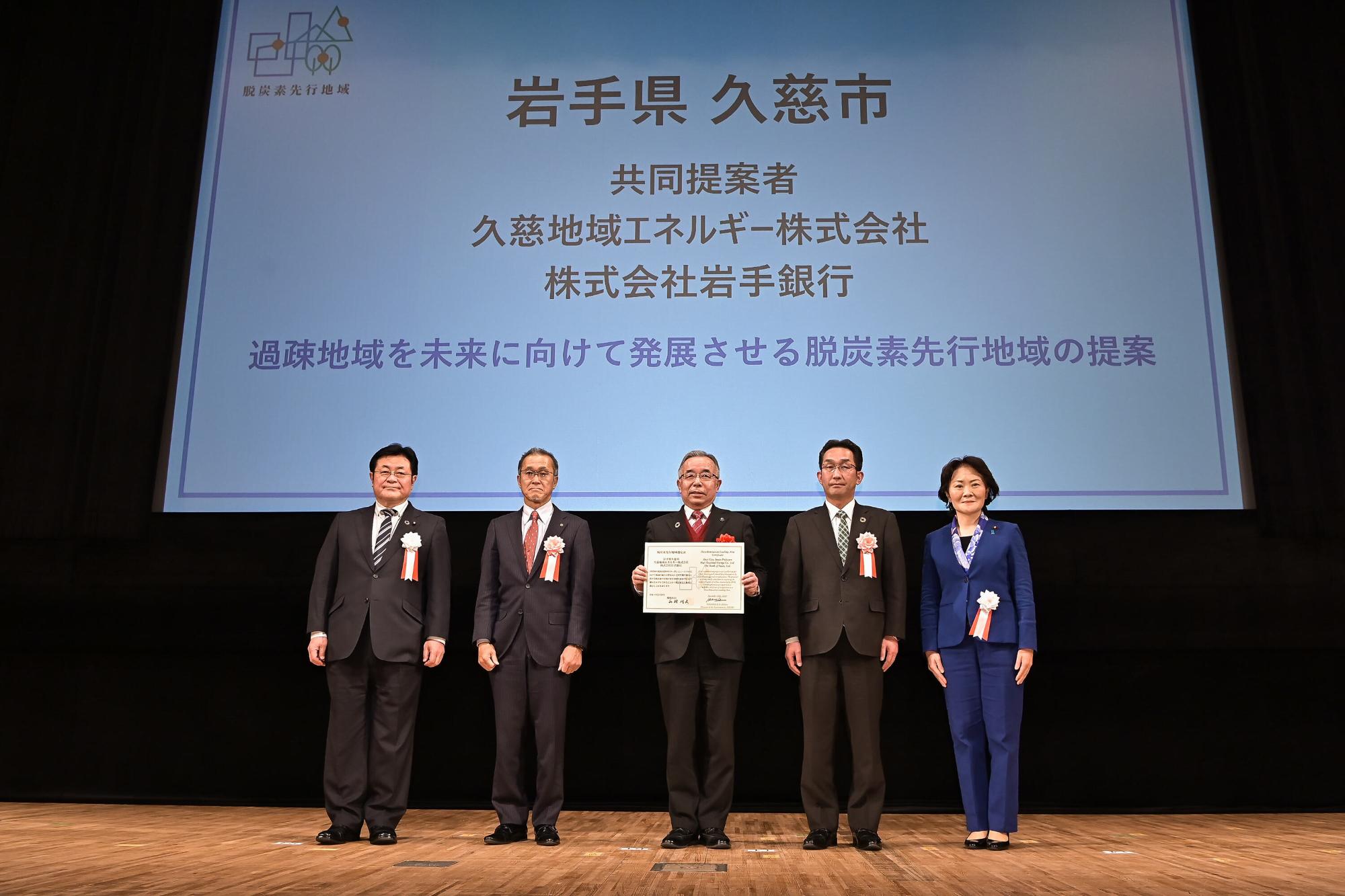 選定書を手に持つ男性を中心にスーツ姿の男女がスクリーンの前で一列に並んでいる写真