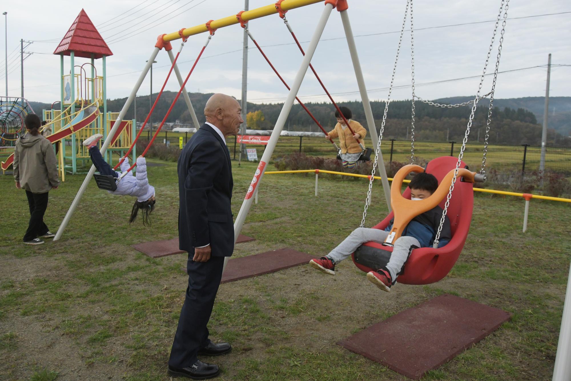 公園のブランコで遊んでいる子供たちをスーツを着た男性が見守っている写真