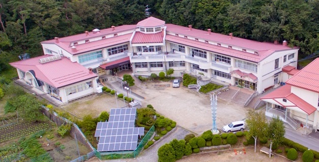 森の前に建つ赤い屋根の校舎とソーラーパネルの置かれた校庭を上空から撮影した写真