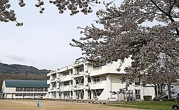 桜の奥に3階建ての校舎と青緑色の屋根の体育館が建っている写真