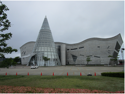 ウェーブ状の屋根の建物の前に円錐の建物が建ってる建物の写真