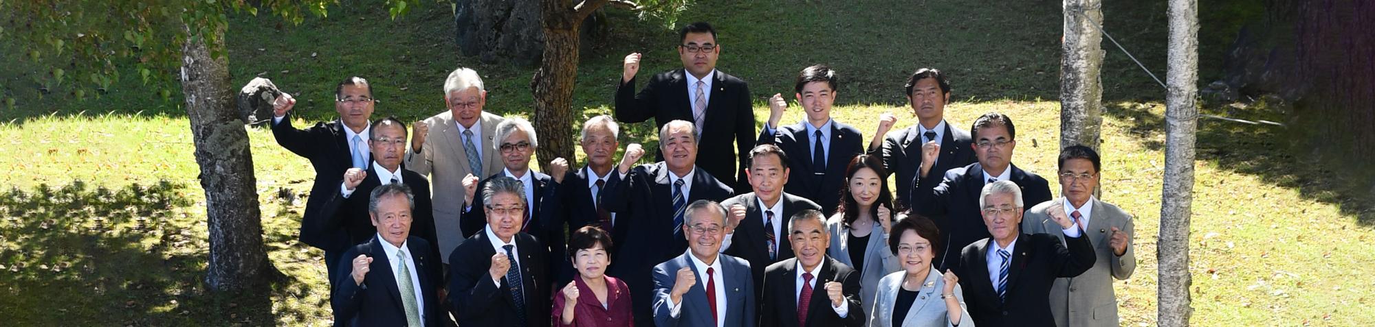 スーツを着た男女20人が木陰でガッツポーズしている写真