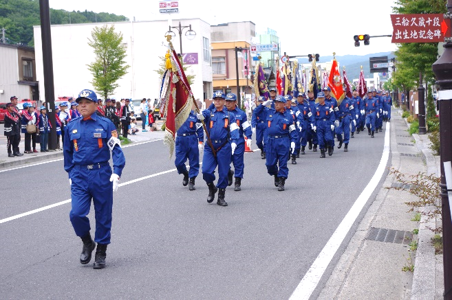 青い活動服姿の消防団員たちが道路を行進している写真