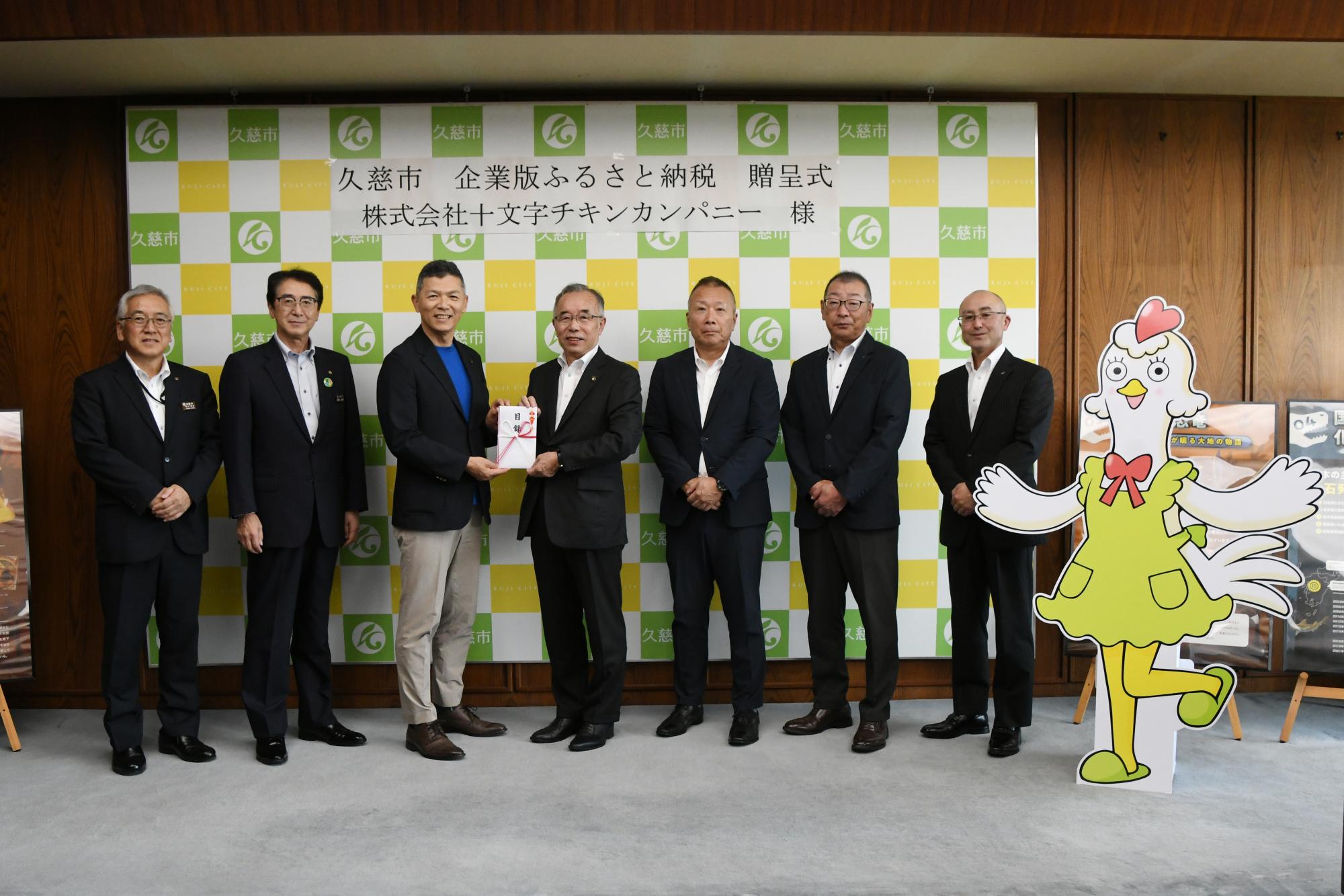 鶏のキャラクターのパネルとスーツ姿の男性たちが一列に並んだ写真