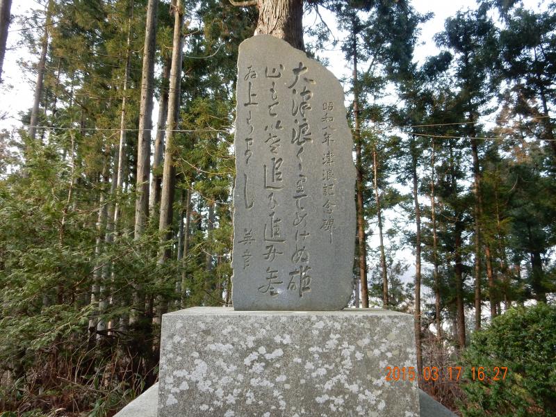 緑生い茂る林を背景に立つ、文字が刻まれた縦長の石碑の写真