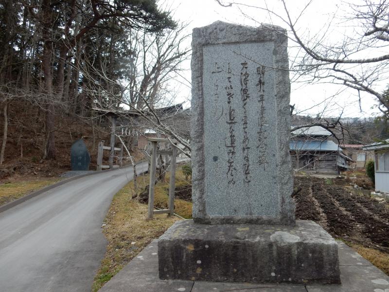 細い道路脇に立つ、文字が刻まれた長方形の石碑の写真