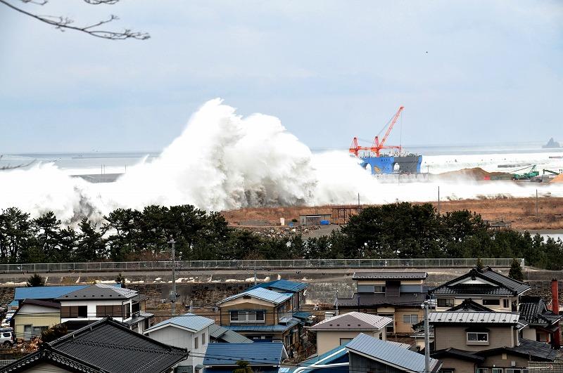 手前に住宅街その後ろに堤防と海岸林があり、さらに奥に大きく高い白い、海しぶきをあげている波の写真