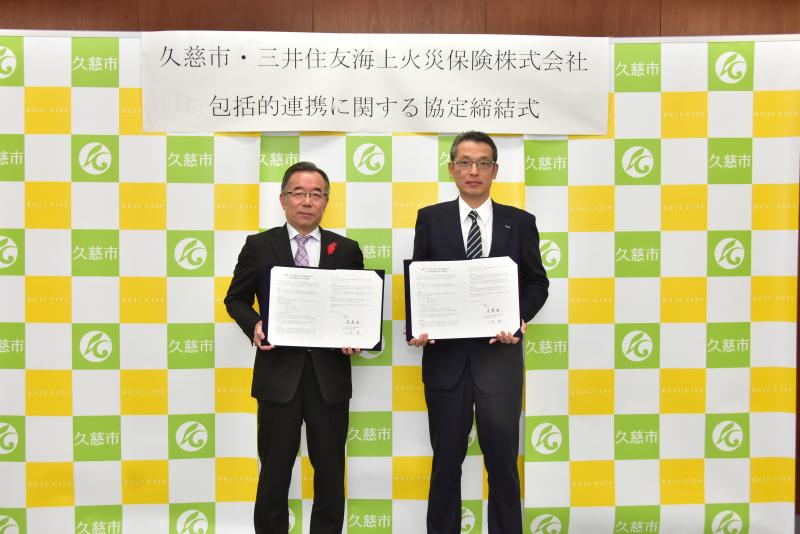 「久慈市・三井住友海上火災保険株式会社 包括的連携に関する協定締結式」と書かれた横断幕を背にスーツ姿の男性2人が証書を見せている写真