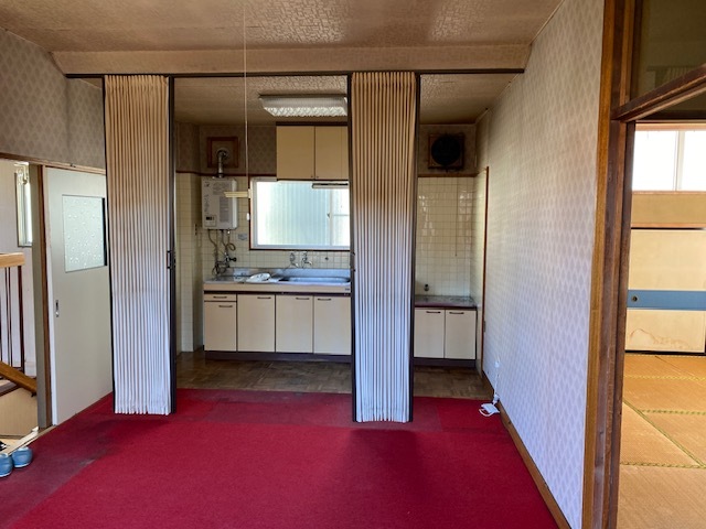 真っ赤な絨毯の敷かれた部屋の奥にカーテンで仕切ることのできるキッチンがある部屋の写真