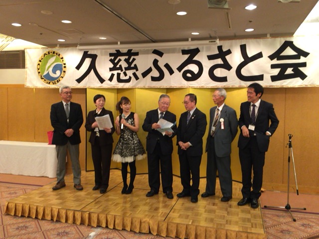 「久慈ふるさと会」と書かれた横断幕の前でスーツやドレスを着た男女が一列に並んでいる写真