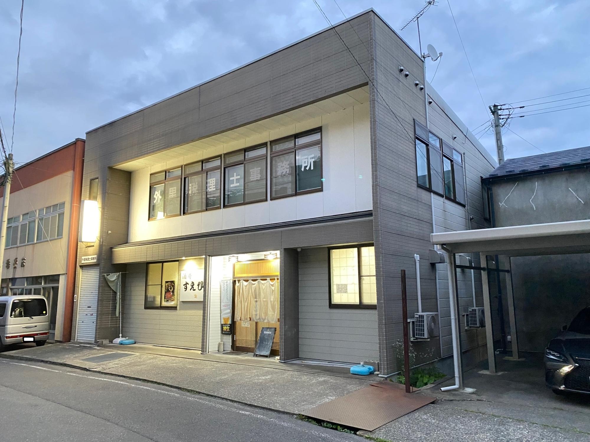 グレーと白色の二階建ての建物の一階の入り口に暖簾がかかっており、明かりがついていている写真